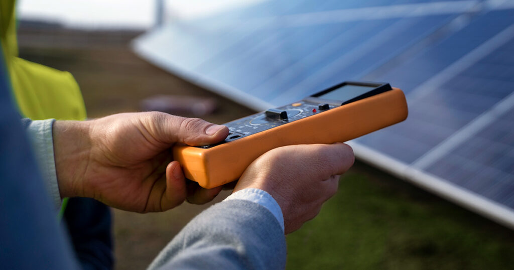 A person holding an electronic device in front of a solar panel, inspecting and measuring its performance.