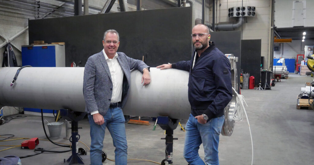 Twee technici poseren voor een stalen buis in een werkplaats, omgeven door gereedschap en machines.